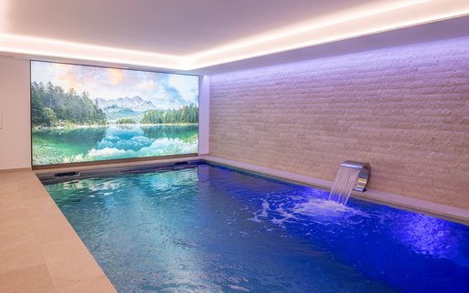 Modernes Hallenbad mit Wasserfall-Einlauf, stimmungsvoller LED-Beleuchtung und großem Natur-Panoramabild an der Wand.