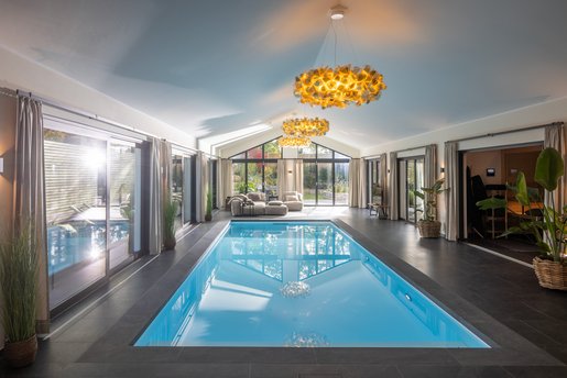 Modern gestaltete Schwimmhalle mit hellem Pool, stilvoller Beleuchtung und gemütlicher Lounge-Ecke vor großen Fensterfronten.