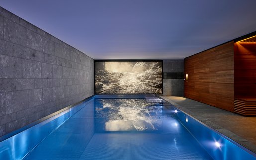 Stimmungsvoll beleuchteter Indoor-Pool mit moderner Beton- und Holzarchitektur sowie großformatigem Wandbild als Blickfang.
