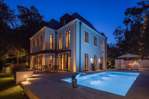 Beleuchteter Außenpool vor stilvoller Villa in der Dämmerung mit Sitzbereich und Garten.