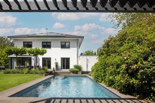 Gepflegter Außenpool vor modernem Einfamilienhaus im Garten, umgeben von üppiger Bepflanzung und überdachter Terrasse.